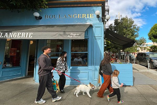 Four people and a dog walk past a blue boulangerie on a sunny day in a city neighborhood.