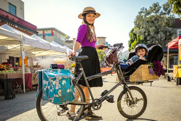 A woman with a hat stands by a bicycle with a child in the back basket at an outdoor market.
