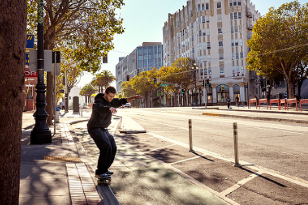 A person skateboards along a city street on a sunny day, with buildings and trees lining the road.