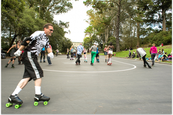 People roller skating on an outdoor rink in a park with trees and others sitting on the grass nearby.