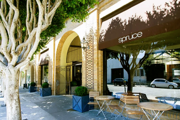 Outdoor cafe tables under trees in front of a building with a brown "Spruce" awning on a sunny day.